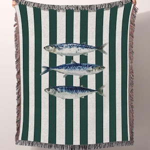 made in the USA. cotton throw blanket with green and white vertical stripes featuring three blue mackerel fish illustrations centered on the design. The rectangular blanket has decorative fringe trim along the top and bottom edges. Someone holds up the blanket to display its full size, showing the textured weave and coastal nautical theme. Perfect for beach houses or coastal-themed home decor, made in the USA.