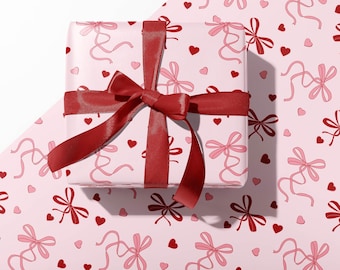 May include: A pink gift box with a red ribbon and bow. The box is wrapped in paper with a pattern of pink bows and red hearts.