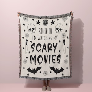 K&ouml;nnte beinhalten: Eine wei&szlig;e gewebte Wolldecke mit schwarzen, gruseligen Halloween-Designs. Die Decke zeigt Sch&auml;del, Flederm&auml;use, Sterne, Spinnweben und Messer. Der Text auf der Decke lautet "Shhh! Ich schaue mir meine Horrorfilme an".