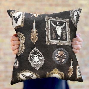 May include: A black and gold throw pillow with a repeating pattern of ornate frames featuring various spooky and gothic imagery, including a skull, a ghost, a bat, a rose, and candles.
