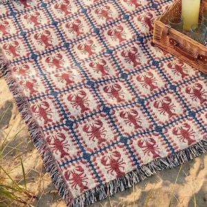 May include: A woven picnic blanket with a repeating pattern of red lobsters on a cream background, framed by a blue and white checkered design. A wicker picnic basket, glasses, and a bottle of lemonade sit on the blanket, perfect for a day at the beach.