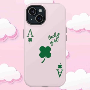 May include: A light pink phone case featuring a green Ace of Clubs card design with a four-leaf clover and the words "lucky girl" in a script font. The case has a black camera lens.