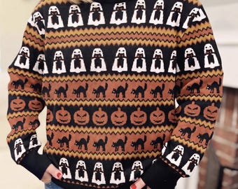 May include: A long-sleeve crewneck sweater featuring a repeating pattern of white ghosts, black arched cats, and orange jack-o'-lanterns on a black and brown background. The knit garment has a ribbed black collar, cuffs, and hemline.