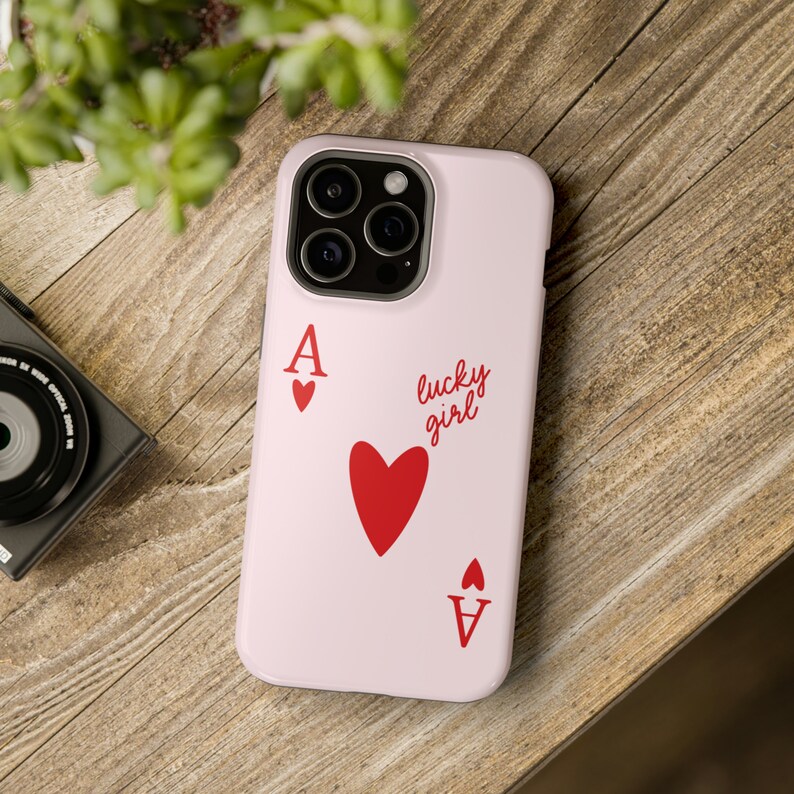 May include: A pink phone case with a red ace of hearts playing card design. The case has a black camera lens and the text "lucky girl" printed on it.