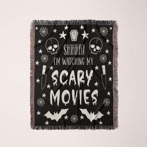 K&ouml;nnte beinhalten: Schwarze und wei&szlig;e gewebte Wurfdecke mit einem gruseligen Halloween-Thema. Sie zeigt Sch&auml;del, Flederm&auml;use, Sterne, Spinnweben und ein Messer. Der Text lautet "Psst! Ich schaue mir meine Horrorfilme an."