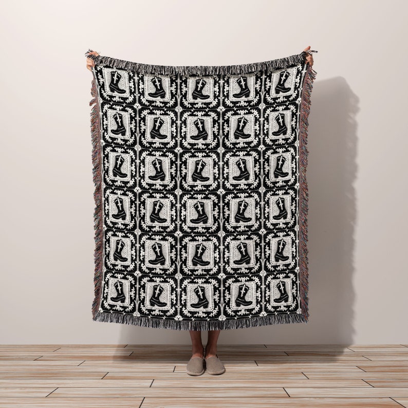 Western-inspired woven throw blanket featuring repeating cowboy boot pattern in black and white. Square grid design with fringed edges, displayed held up against neutral wall. Soft textile with rustic charm, perfect for country or ranch decor