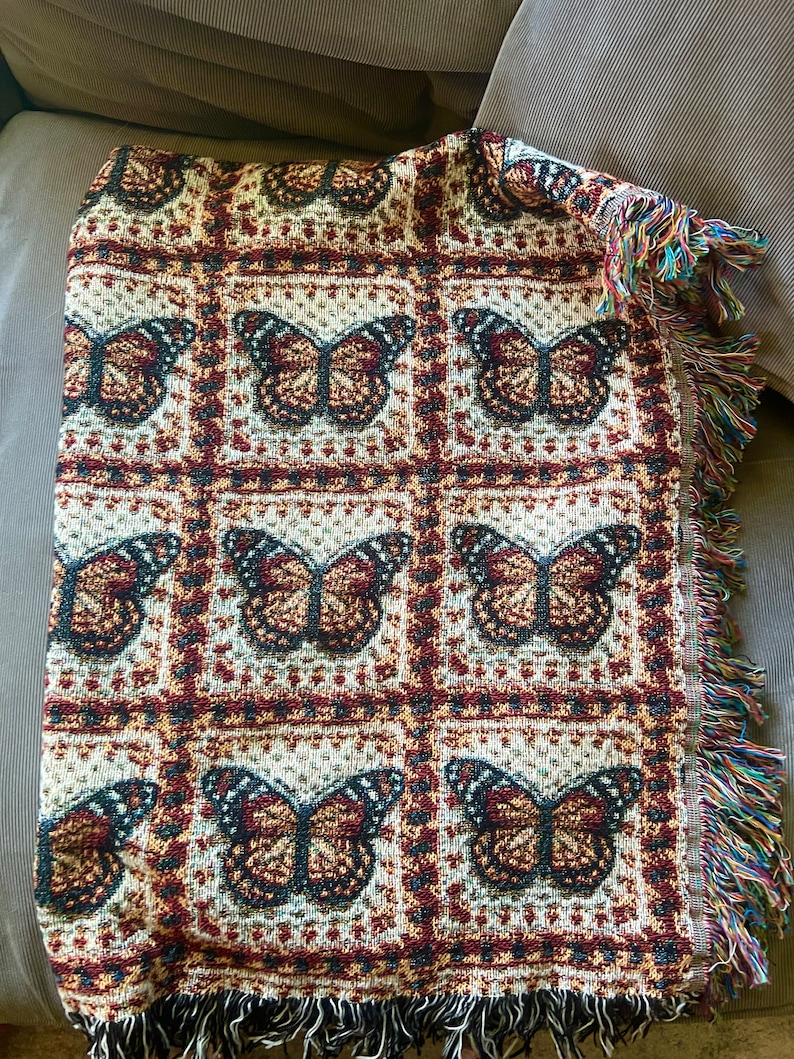 May include: Woven throw blanket with a repeating butterfly pattern in brown, red, and cream. The blanket has a fringed edge with multiple colours. The butterfly design is set within a grid pattern.