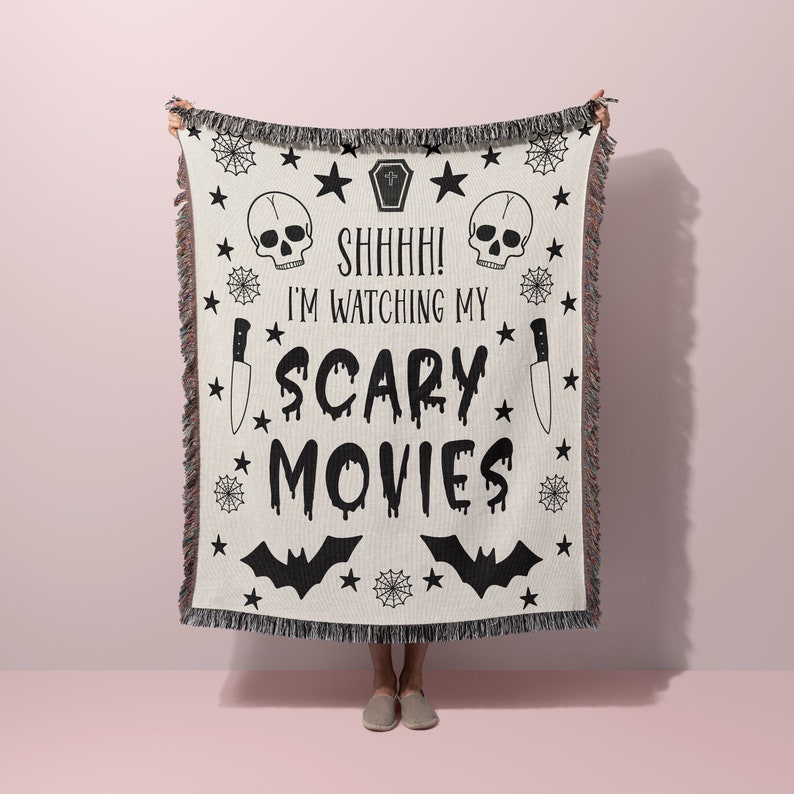 K&ouml;nnte beinhalten: Eine wei&szlig;e gewebte Wolldecke mit schwarzen, gruseligen Halloween-Designs. Die Decke zeigt Sch&auml;del, Flederm&auml;use, Sterne, Spinnweben und Messer. Der Text auf der Decke lautet "Shhh! Ich schaue mir meine Horrorfilme an".