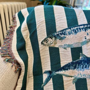 May include: A woven throw blanket with a striped pattern in teal and off-white. The blanket features detailed illustrations of fish in shades of blue and white. The edge of the blanket has a fringe in multiple colors.