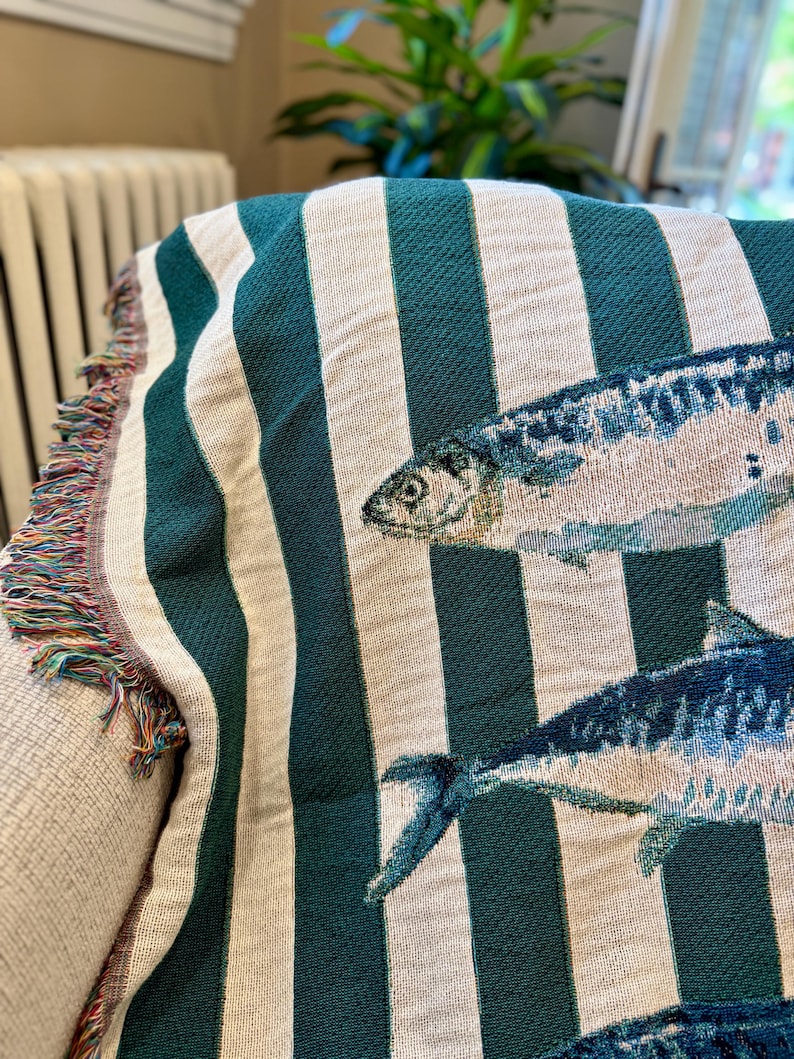 May include: A woven throw blanket with a striped pattern in teal and off-white. The blanket features detailed illustrations of fish in shades of blue and white. The edge of the blanket has a fringe in multiple colors.