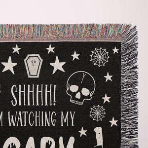 K&ouml;nnte beinhalten: Schwarze gewebte Wurfdecke mit einem gruseligen Halloween-Design. Sie zeigt zwei wei&szlig;e Sch&auml;del, einen Sarg, Spinnweben, Sterne und den Text "Psst! Ich beobachte mein gruseliges..."