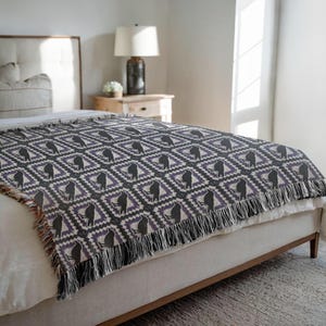 May include: A woven throw blanket with a repeating pattern of black figures within diamond shapes, set against a purple and gray background. The blanket has a fringe along the bottom edge and is draped over a bed.