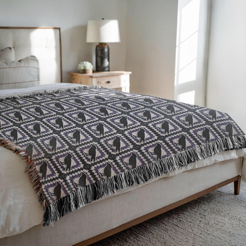 May include: A woven throw blanket with a repeating pattern of black figures within diamond shapes, set against a purple and gray background. The blanket has a fringe along the bottom edge and is draped over a bed.