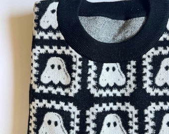 May include: A black and white knit sweater featuring a repeating pattern of white ghost figures enclosed within individual square frames. The garment has a solid black ribbed crew neckline and a consistent grid-like design across the front surface.