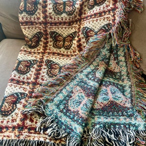 May include: Woven throw blanket with a butterfly design in brown, cream, and burgundy. It has a fringed edge with multicoloured threads and a reversible design. The reverse side features a teal and burgundy pattern.