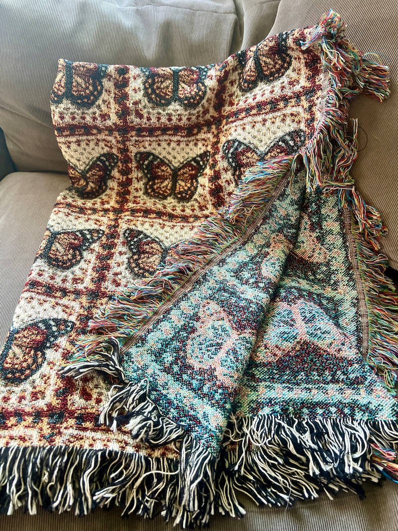 May include: Woven throw blanket with a butterfly design in brown, cream, and burgundy. It has a fringed edge with multicoloured threads and a reversible design. The reverse side features a teal and burgundy pattern.