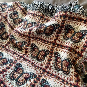 May include: A woven throw blanket with a repeating butterfly design in brown, cream, and burgundy. The blanket has a fringed edge with black and white threads. The design is detailed and the overall aesthetic is cosy.