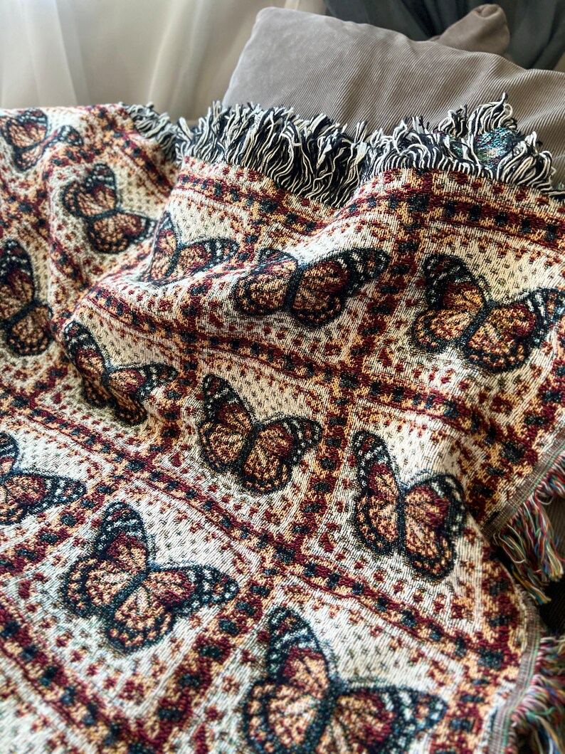 May include: A woven throw blanket with a repeating butterfly design in brown, cream, and burgundy. The blanket has a fringed edge with black and white threads. The design is detailed and the overall aesthetic is cosy.