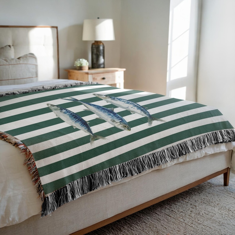 May include: A green and white striped throw blanket with a repeating pattern of three fish. The blanket has a fringed edge and is draped over a bed. The fish are blue and white.