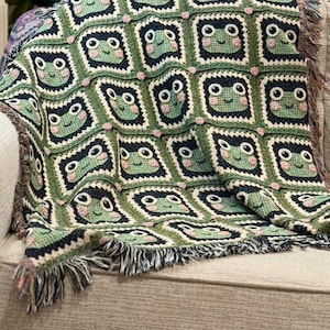 May include: A green and black crocheted throw blanket with a repeating pattern of frog faces. The blanket has fringe on all sides.