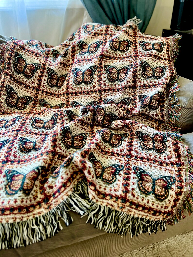 May include: A woven throw blanket with a repeating butterfly pattern in brown, cream, and burgundy. The blanket has a fringed edge with multiple colours. The blanket is draped over a grey sofa.