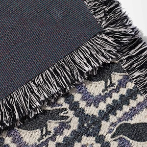 May include: Woven throw blanket with a geometric pattern in shades of black, white, and purple. The blanket features a fringe border in black and white. The underside of the blanket is a solid dark teal color.