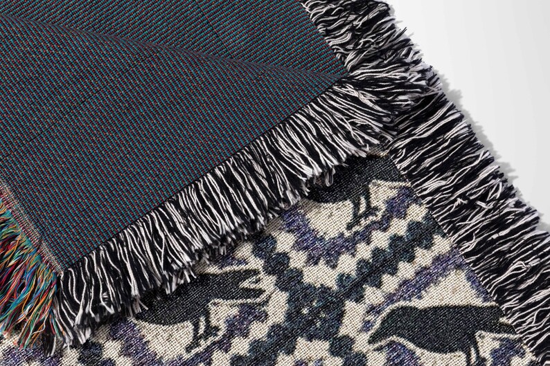 May include: Woven throw blanket with a geometric pattern in shades of black, white, and purple. The blanket features a fringe border in black and white. The underside of the blanket is a solid dark teal color.