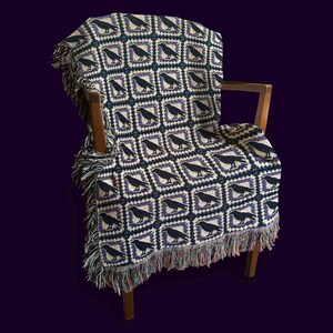 May include: A woven throw blanket draped over a wooden armchair. The blanket features a repeating pattern of black crows within square frames, set against a cream and purple background. The edges are finished with a fringed trim.
