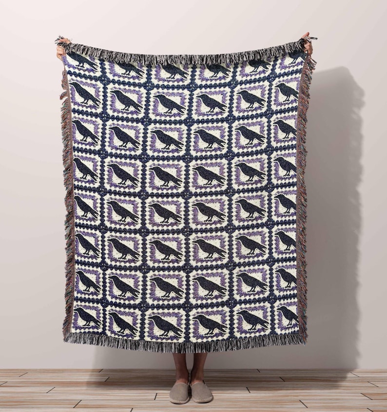 Woman holding up a cotton woven throw tapestry blanket with a purple and black and white granny square with a raven crow in the center.