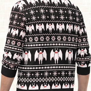 May include: A black long-sleeve sweater featuring a repeating white pixelated moth pattern with red eyes, interspersed with white geometric snowflake-like designs and horizontal stripes, finished with a ribbed crew neckline and cuffs.