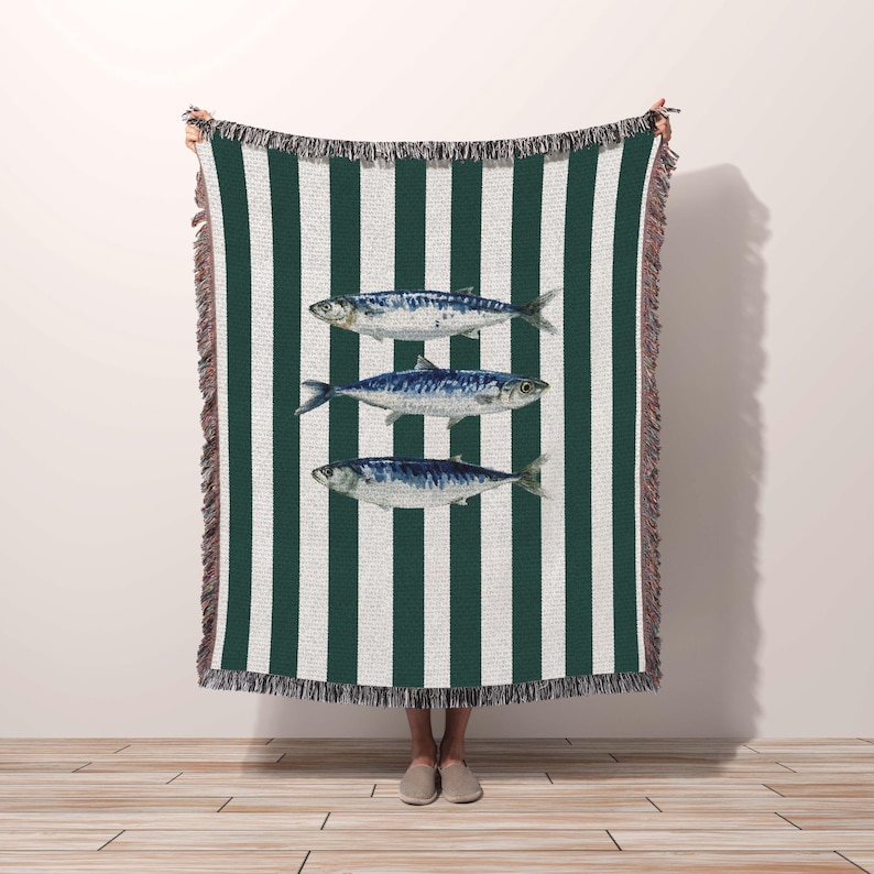 made in the USA. cotton throw blanket with green and white vertical stripes featuring three blue mackerel fish illustrations centered on the design. The rectangular blanket has decorative fringe trim along the top and bottom edges. Someone holds up the blanket to display its full size, showing the textured weave and coastal nautical theme. Perfect for beach houses or coastal-themed home decor, made in the USA.