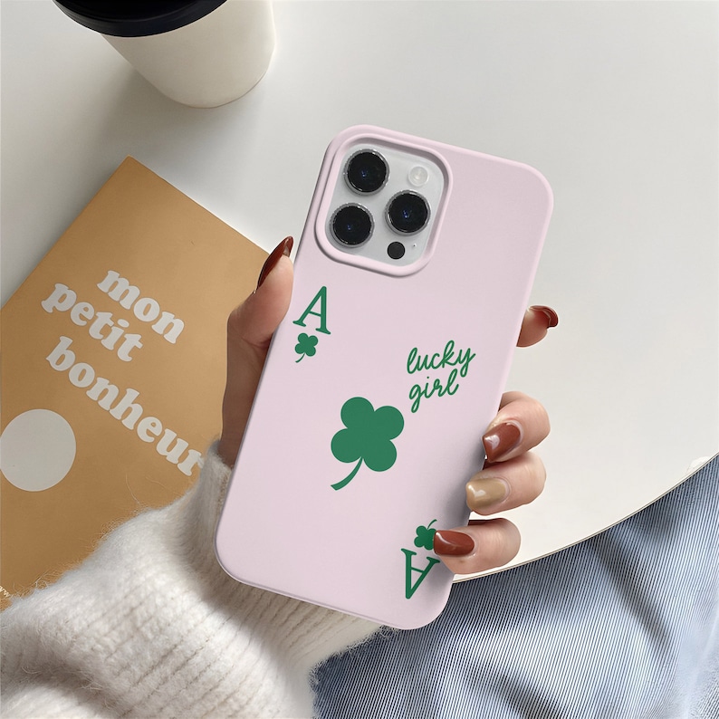 May include: A light pink phone case featuring a green four-leaf clover and the text "lucky girl". The design includes the Ace of Clubs playing card symbol. The phone case is shown with a white cup and a book.