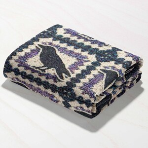 May include: Folded blanket featuring a black raven silhouette on a cream background with a geometric pattern in shades of purple and navy blue. The blanket is folded neatly, showcasing the design.