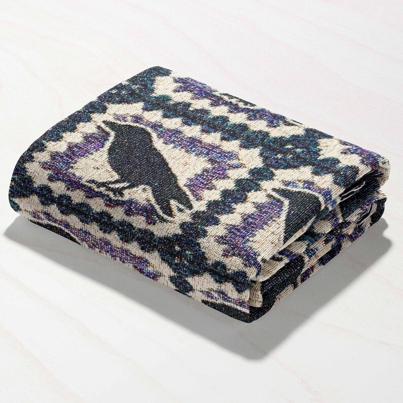 May include: Folded blanket featuring a black raven silhouette on a cream background with a geometric pattern in shades of purple and navy blue. The blanket is folded neatly, showcasing the design.