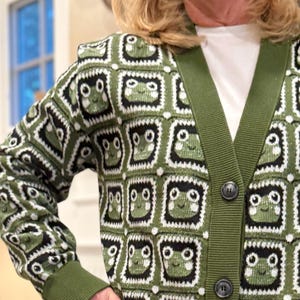 May include: A green cardigan with a white and black frog pattern. The cardigan has a button closure and long sleeves.