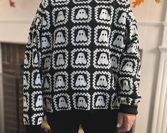 May include: A long-sleeved sweater featuring a repeating grid pattern of white ghost motifs on a black background. The garment has a crew neckline, ribbed cuffs, and a ribbed hem, constructed with a knit texture that creates a square block design.