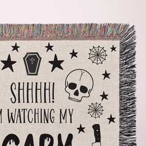 K&ouml;nnte beinhalten: Eine gewebte Wandbehang mit einem schwarz-wei&szlig;en Design mit Totenk&ouml;pfen, Sternen, einem Sarg, Spinnweben und einem Messer. Der Text "Shhh! I'm watching my scary" ist auf dem Wandbehang gedruckt.