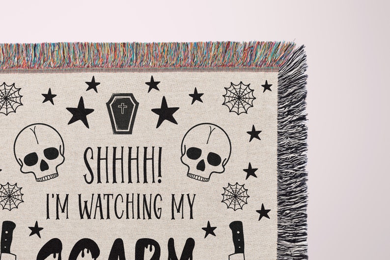 K&ouml;nnte beinhalten: Eine gewebte Wandbehang mit einem schwarz-wei&szlig;en Design mit Totenk&ouml;pfen, Sternen, einem Sarg, Spinnweben und einem Messer. Der Text "Shhh! I'm watching my scary" ist auf dem Wandbehang gedruckt.