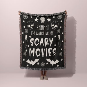 K&ouml;nnte beinhalten: Schwarze und wei&szlig;e gewebte Wolldecke mit einem gruseligen Halloween-Motiv. Sie zeigt einen Totenkopf, Flederm&auml;use, Sterne, Spinnweben und den Text "Psst! Ich schaue mir meine Horrorfilme an".