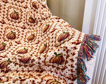 May include: A woven throw blanket with a white background and a pattern of orange and brown pumpkins. The blanket has a fringe border with multi-coloured threads.