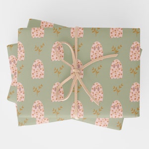 Pode incluir: Uma pilha de papel de embrulho verde com um padr&atilde;o repetitivo de fantasmas rosa com desenhos florais laranja e branco. O papel &eacute; amarrado com uma fita marrom.