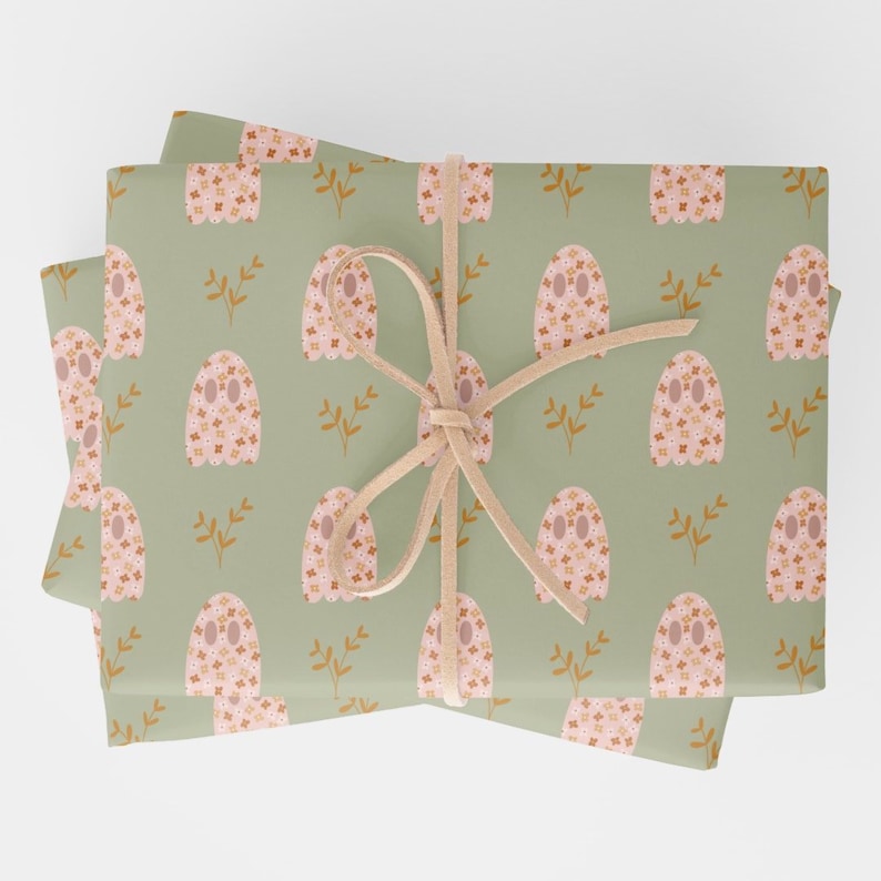 Pode incluir: Uma pilha de papel de embrulho verde com um padr&atilde;o repetitivo de fantasmas rosa com desenhos florais laranja e branco. O papel &eacute; amarrado com uma fita marrom.