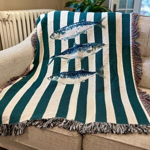 May include: A woven throw blanket with a striped pattern in teal and white, draped over a sofa. The blanket features three detailed illustrations of fish in shades of blue and white. The edges are finished with a fringed trim.