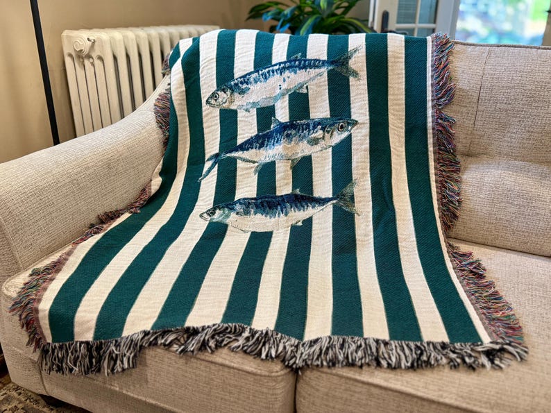 May include: A woven throw blanket with a striped pattern in teal and white, draped over a sofa. The blanket features three detailed illustrations of fish in shades of blue and white. The edges are finished with a fringed trim.