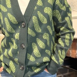May include: Dark green cardigan sweater with a repeating pattern of light green pickles. The sweater has a V-neck, dark green trim, and three black buttons. The person is wearing blue jeans and a white t-shirt underneath.