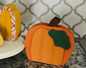May include: A set of five wooden pumpkins in various colors and patterns, including red, orange, yellow, and plaid. The pumpkins are arranged on a white cake stand. A wooden leaf with the word "Fall" is also included.