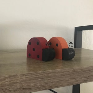Könnte beinhalten: Zwei hölzerne Marienkäfer-Figuren mit schwarzen Punkten und schwarzen bemalten Köpfen. Die Marienkäfer sind in Rot- und Orangetönen bemalt.