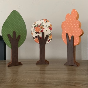 Puede incluir: Tres árboles de madera recortados con diferentes hojas de papel estampado. El árbol de la izquierda tiene hojas verdes con lunares blancos. El árbol del medio tiene hojas con estampado floral en tonos rojos, naranjas y amarillos. El árbol de la derecha tiene hojas naranjas con un patrón de trama blanca.