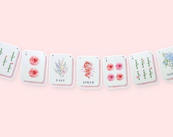 May include: A decorative garland featuring seven rectangular cards with rounded corners, each displaying watercolor illustrations of flowers, bamboo stalks, a red dragon, and the text labels EAST, JOKER, and NORTH, all strung together on a thin cord.