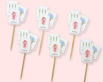 May include: A set of five decorative food picks, each featuring a rectangular card design with a red dragon illustration, the word JOKER, a green bamboo graphic, and a blue hydrangea flower. Each card is attached to a thin, pointed wooden stick.